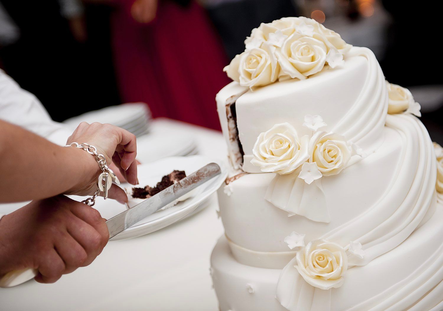 Hands cutting into a tiered wedding cake with white roses.