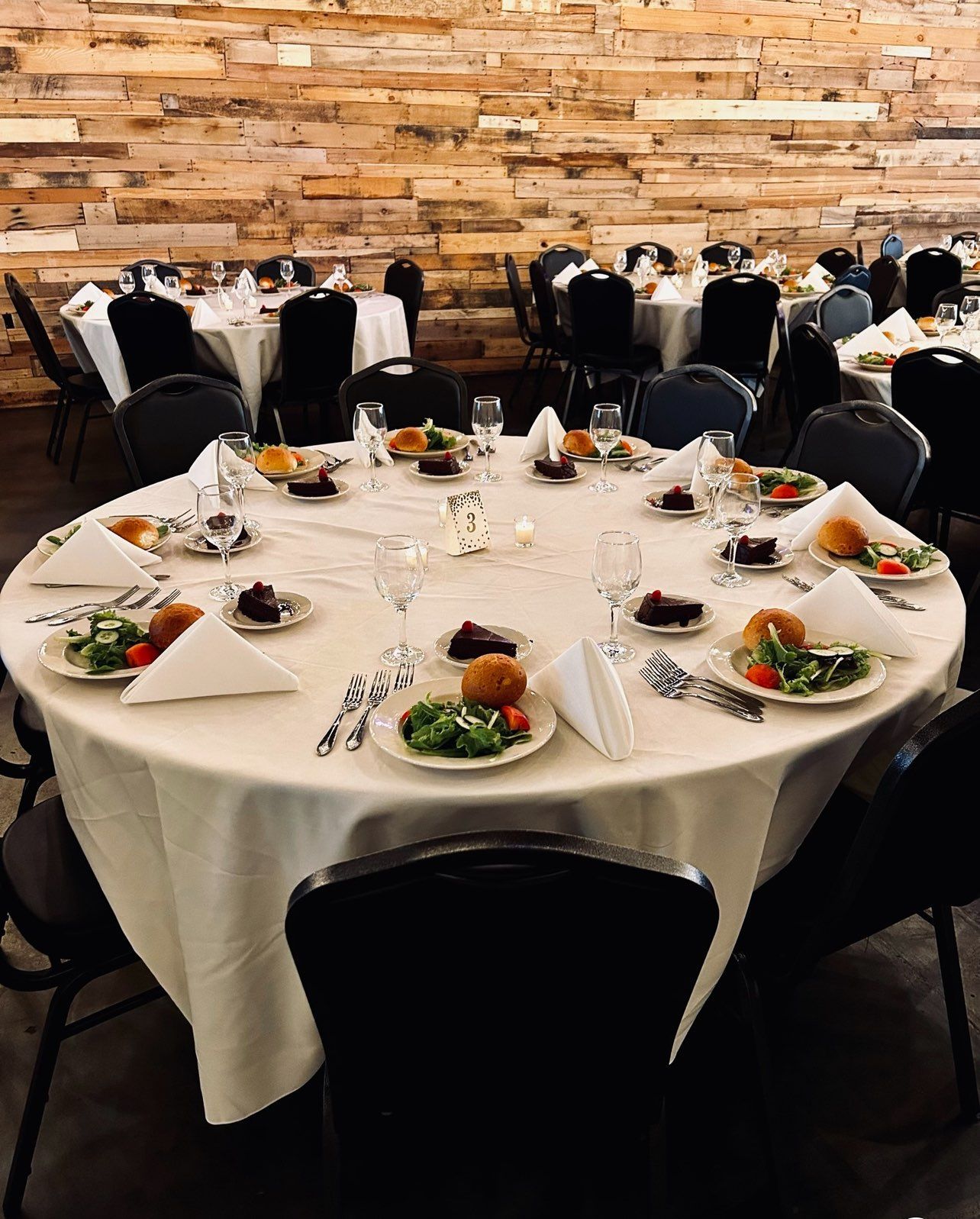 Round tables set for a banquet with white linens, place settings, and food; black chairs in a room with a wooden plank wall.