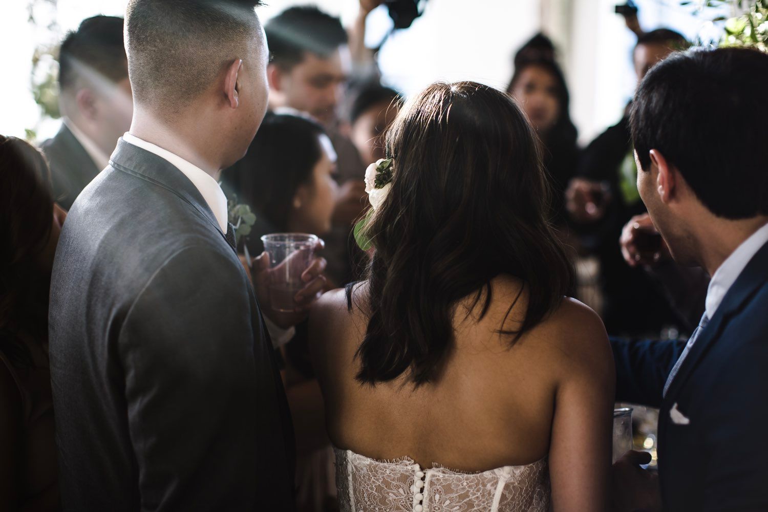 Bride in a wedding dress, surrounded by guests with drinks, smiling and talking at an outdoor reception.
