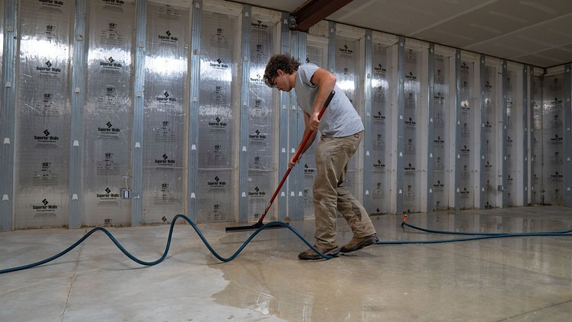 A person in a gray shirt and tan pants uses a floor squeegee to push water across a concrete floor in a room under renovation.