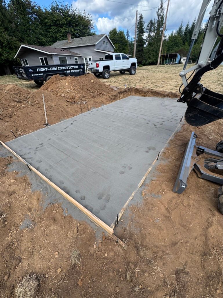 Freshly poured concrete pad in a dirt lot; excavator arm in the frame; background of house, truck, trees.