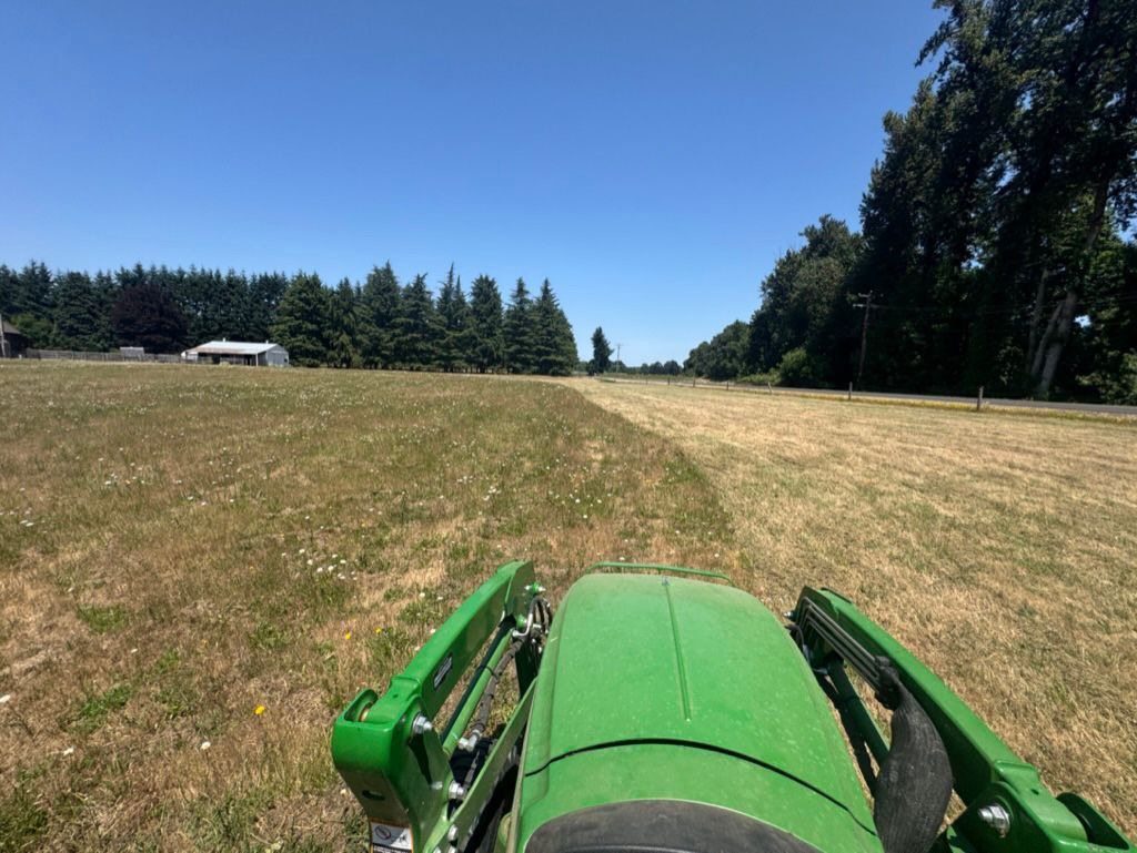 Tractor mowing dry grassy field on a sunny day, with trees and a building in the background.