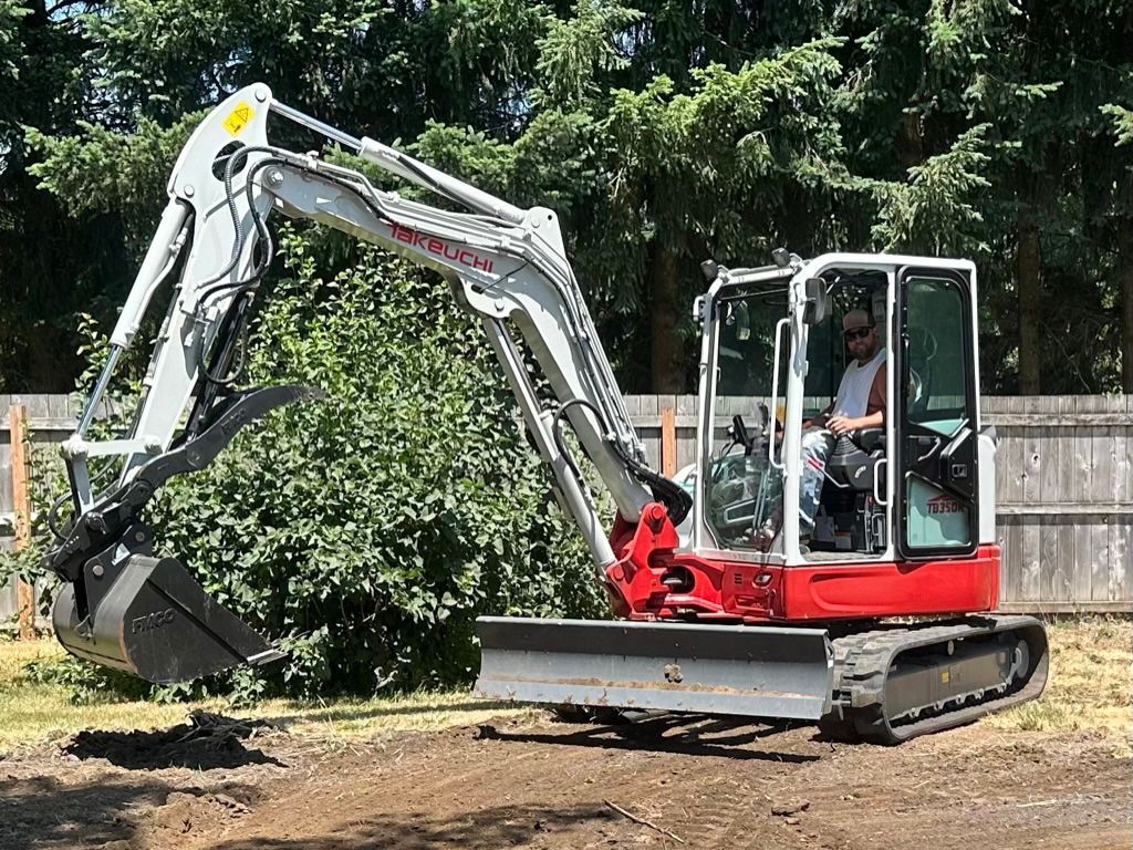 Compact red and white excavator digging in a grassy yard; a person is in the operator's seat.