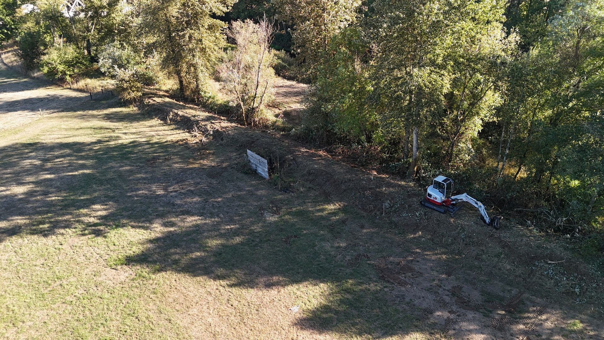 An excavator works beside a treeline, in a grassy area under a blue sky.