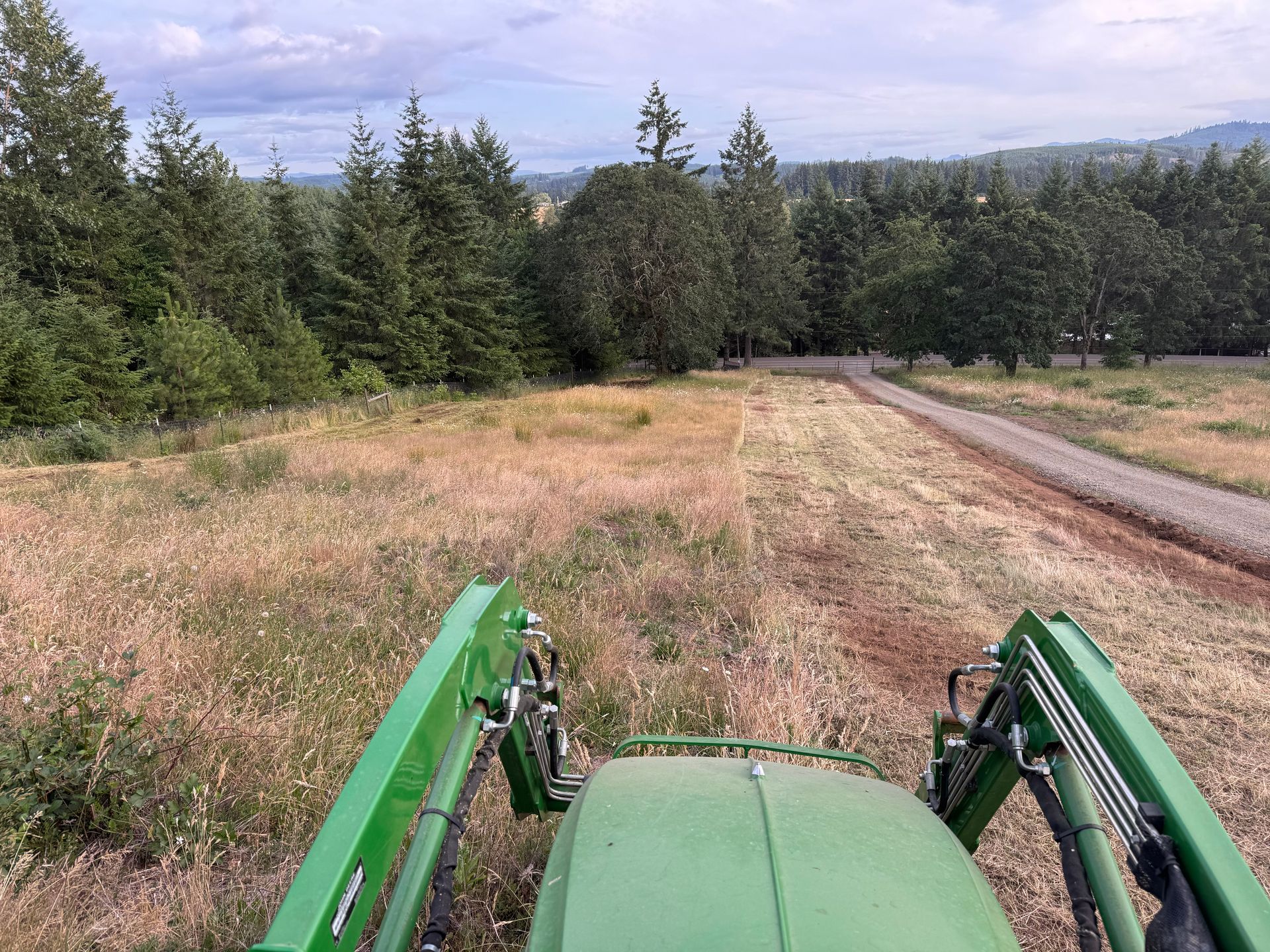 Green tractor mowing a field, forest in the background, partly cloudy sky.
