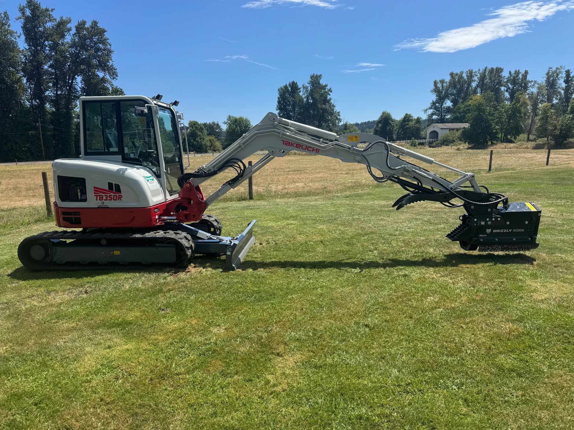 Mini excavator on grass with a brush cutter attachment under a blue sky.