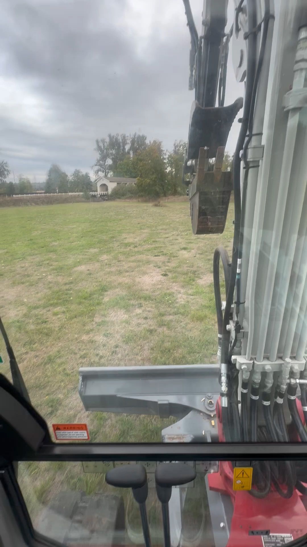 View from an excavator cab overlooking a green field and distant house on an overcast day.