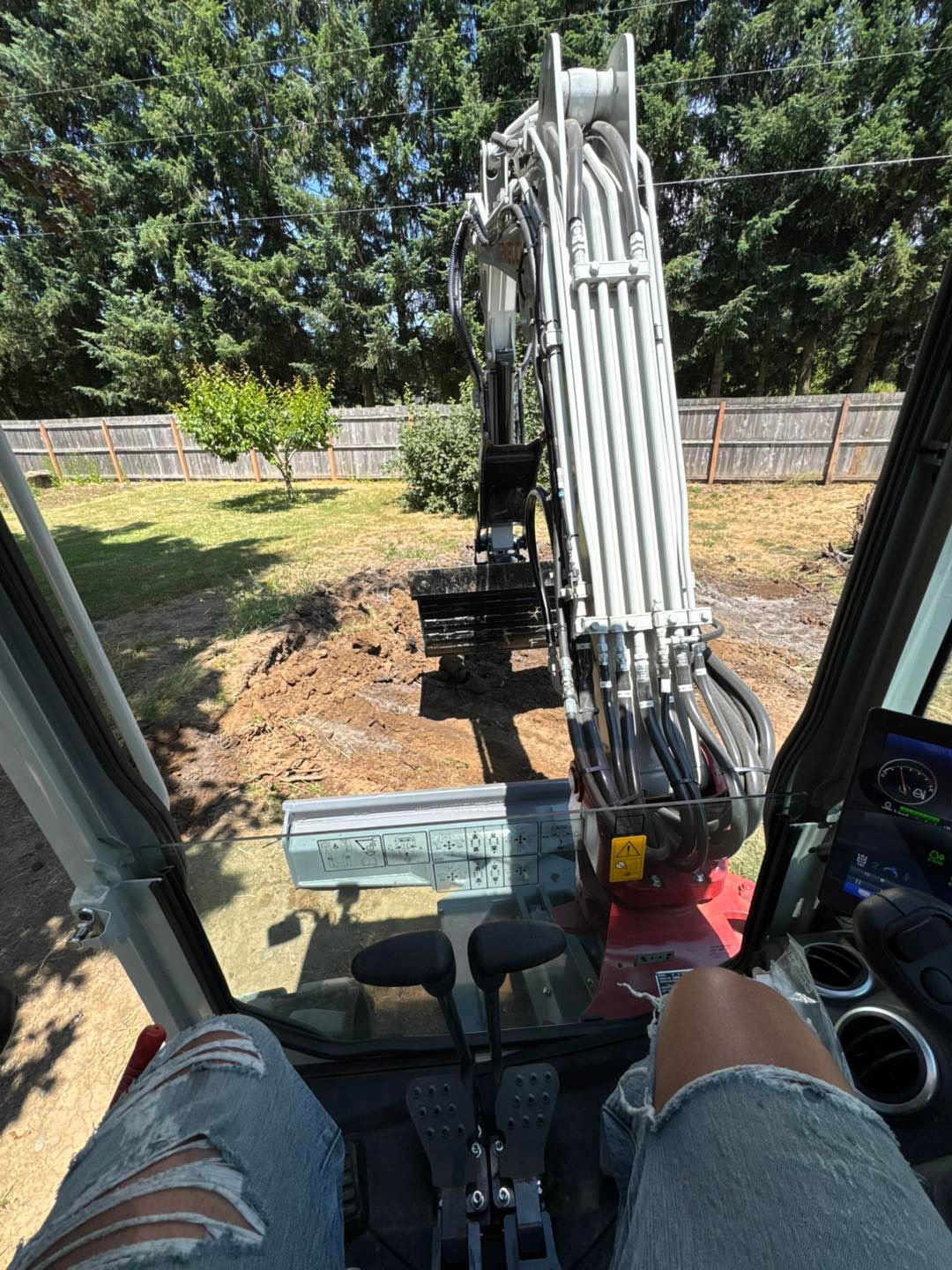 View from inside an excavator, operating arms and pedals, digging in a backyard on a sunny day.