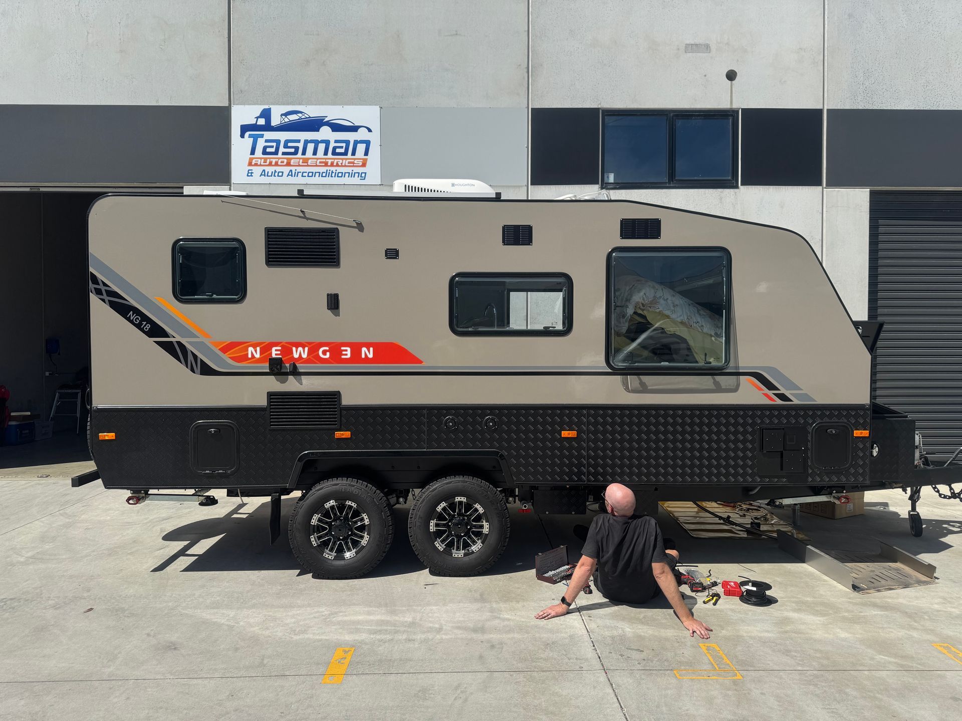Tan and black caravan with person working on it outside a building.