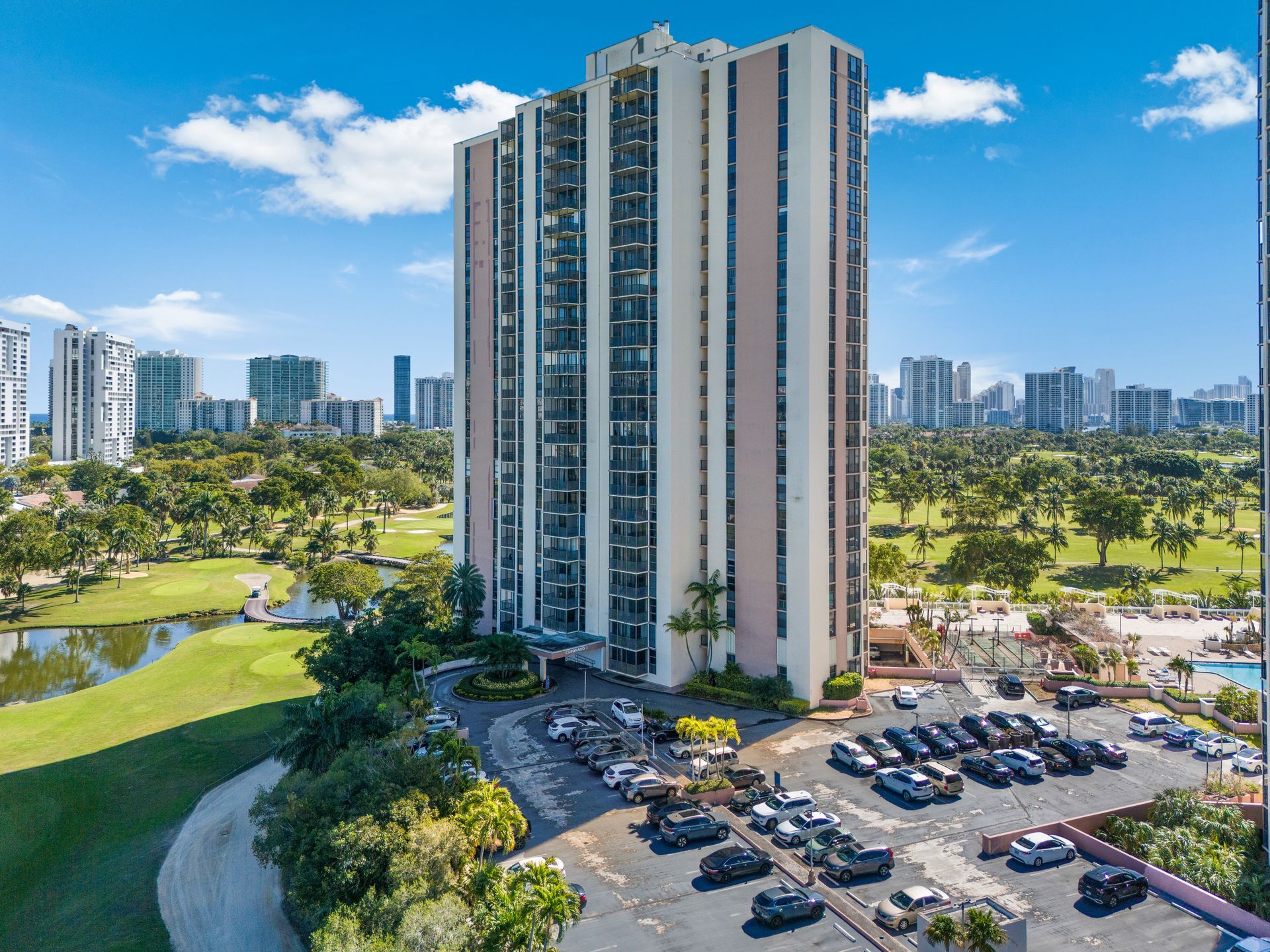 An aerial view of a tall residential tower surrounded by a golf course, greenery, and a parking lot on a sunny day.