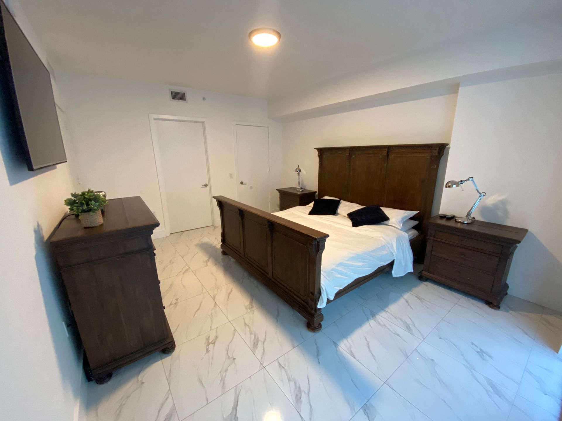 A bedroom with dark wood furniture, including a bed, dresser, and nightstands, against a white wall on marble-tiled floors.