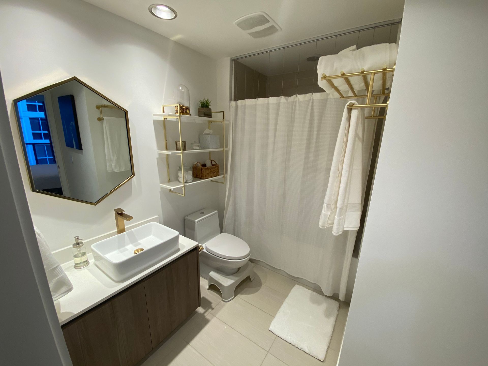 A modern bathroom featuring a white vanity with a gold faucet, a hexagonal mirror, wall shelving, and a shower curtain.