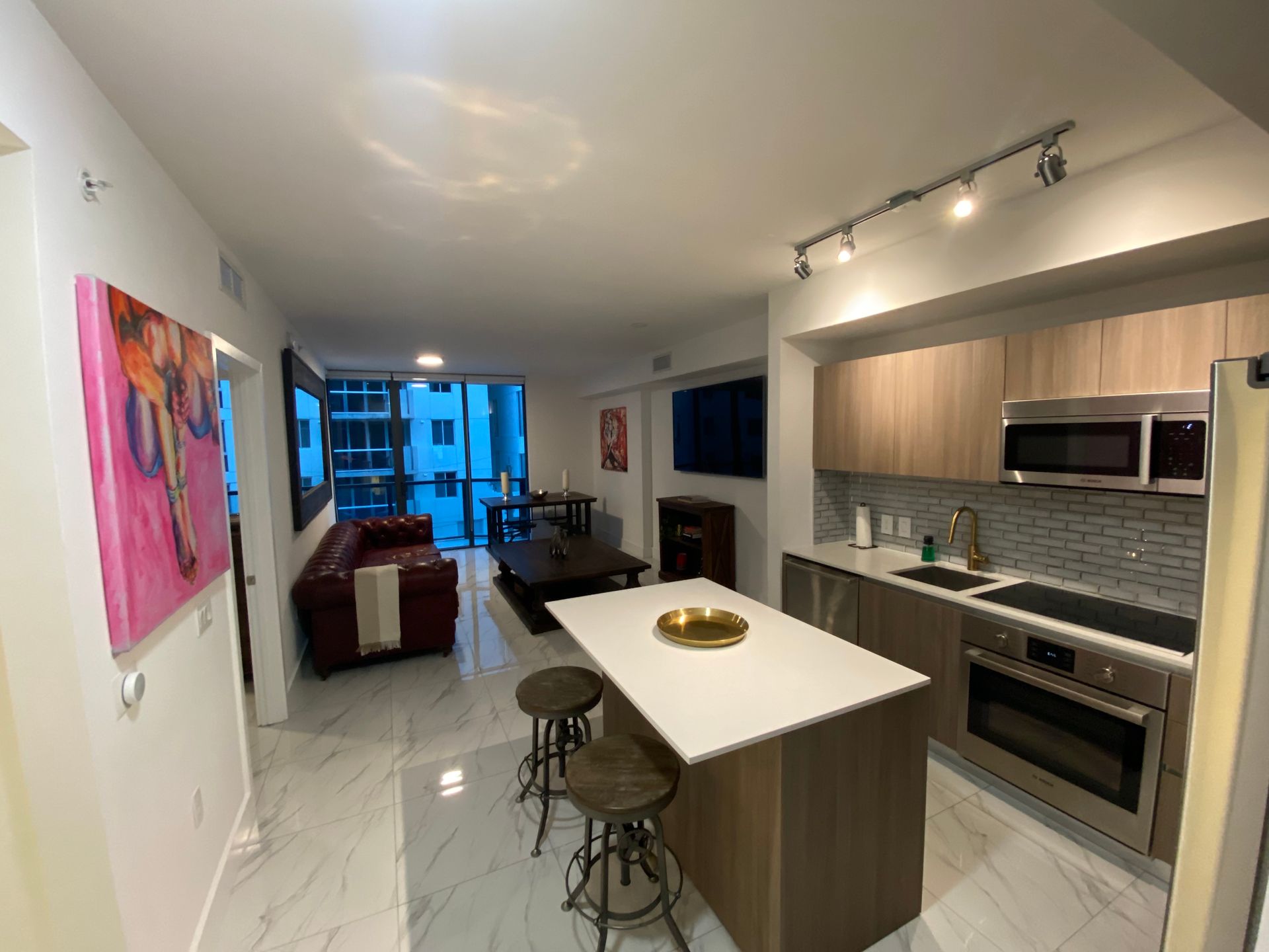 An open-concept condo with a kitchen island and bar stools in the foreground, leading to a living area with a red sofa.