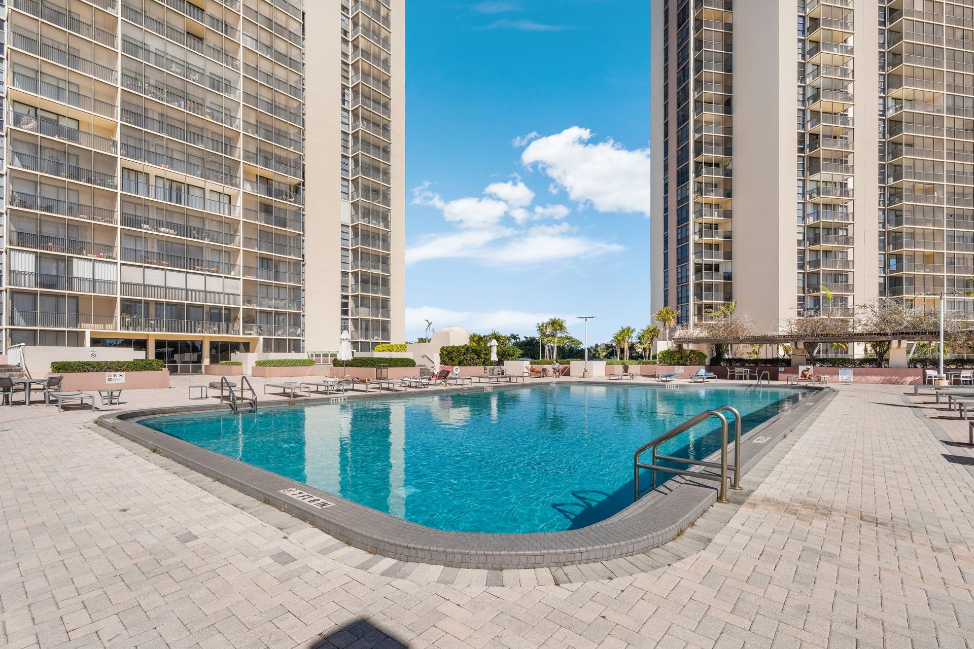 A bright blue swimming pool sits on a paved patio between two high-rise apartment buildings under a sunny sky.