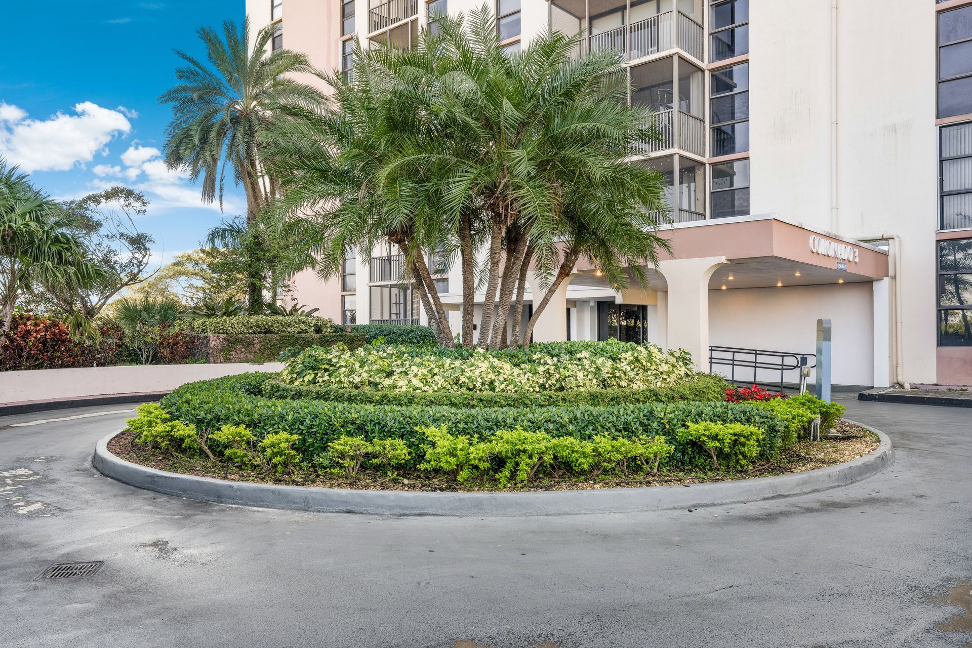 A paved circular driveway with a landscaped garden and palm tree in front of a multi-story building entrance.