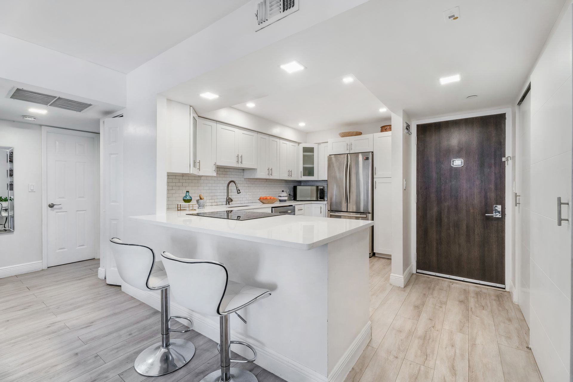 A modern kitchen with white cabinetry, stainless steel appliances, and a breakfast bar with two white stools.