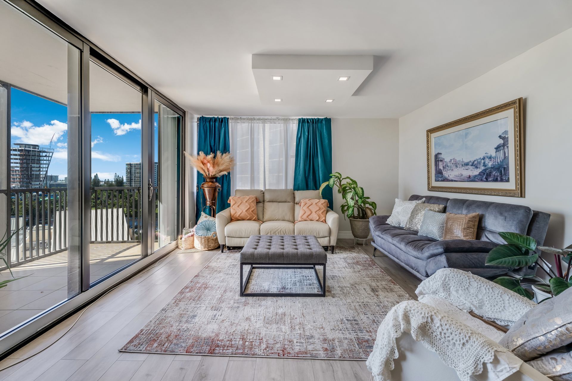 A modern living room with a gray sofa, light beige couch, patterned rug, square ottoman, and floor-to-ceiling windows.