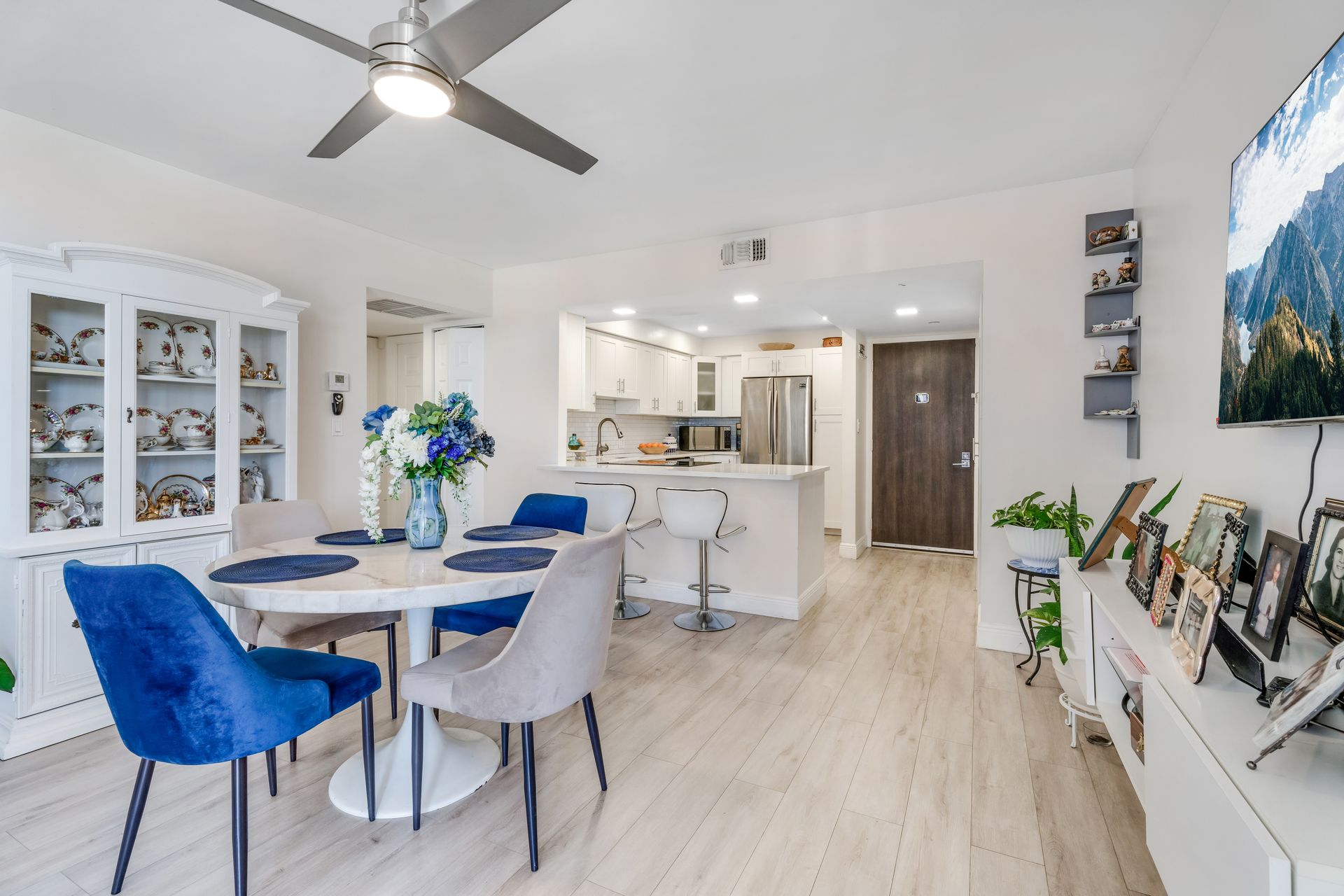 A dining area with a round table, a blue velvet chair, light gray chairs, a white china cabinet, and an open kitchen.