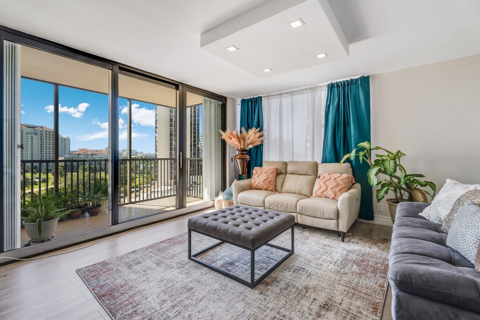 A living room with a beige sofa, gray ottoman, and dark blue curtains, featuring large glass doors opening to a balcony.