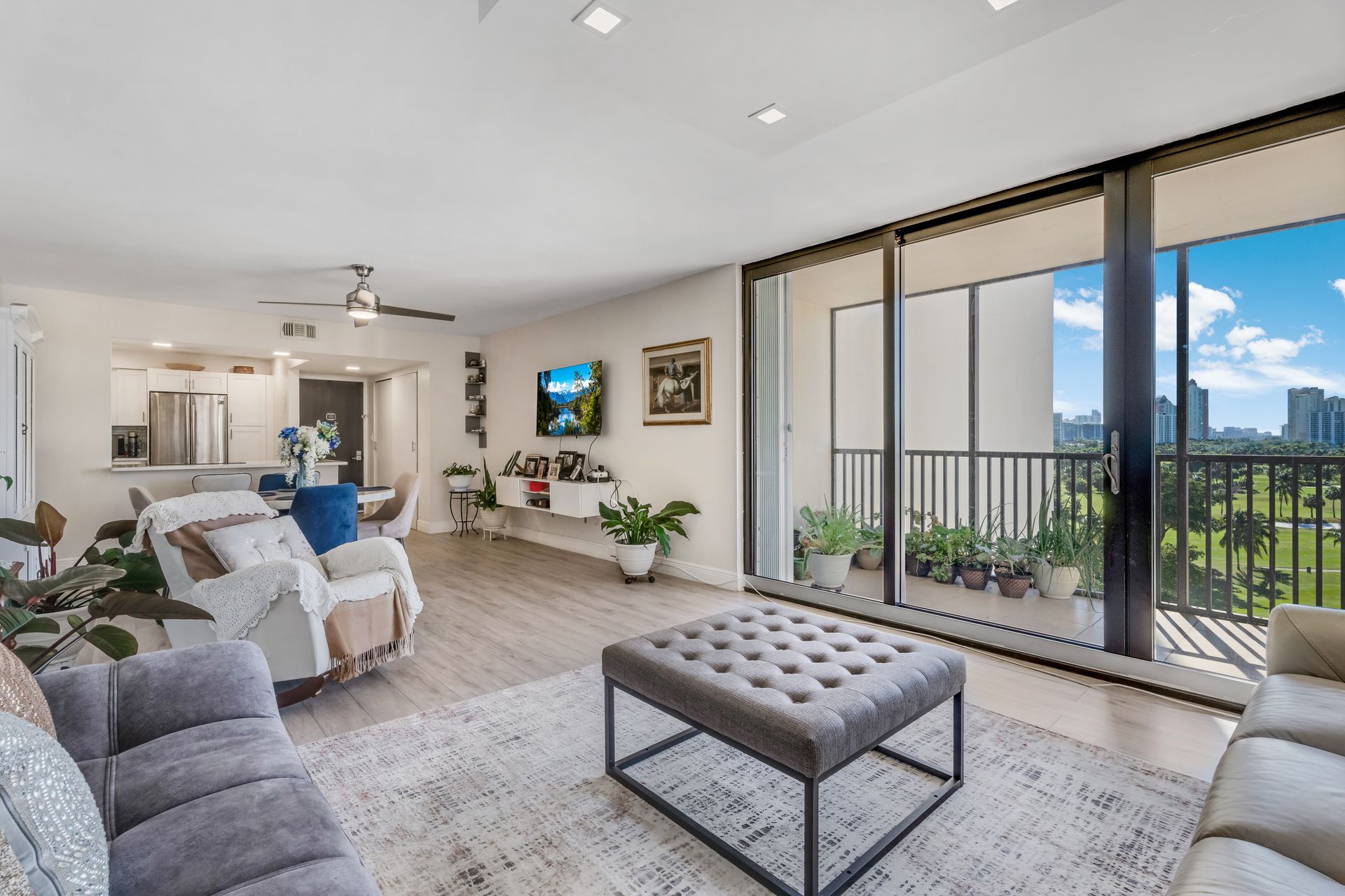 A spacious, modern apartment living room with a grey tufted ottoman, white armchair, and balcony views of city buildings.