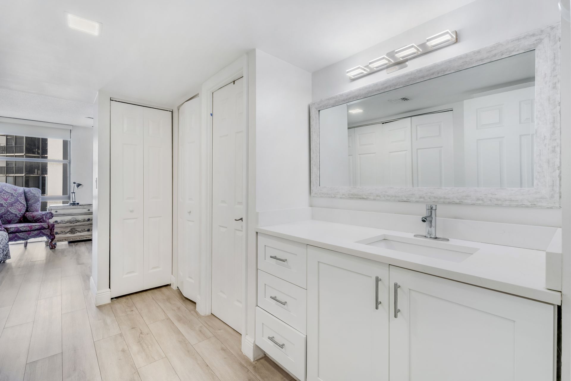 A bright bathroom featuring a white vanity with a single sink, large mirror, and closed closet doors on a light wood floor.
