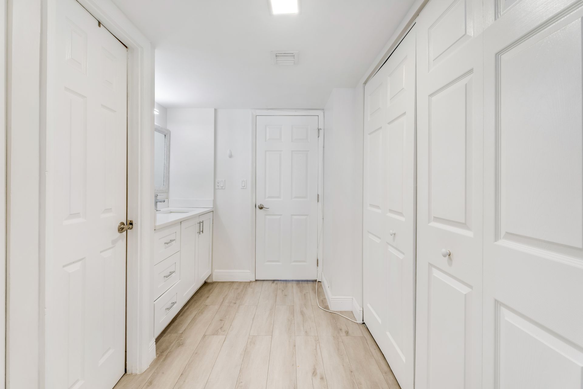 A hallway with light wood-look flooring, white paneled doors on both sides, and a view into a bright white bathroom.