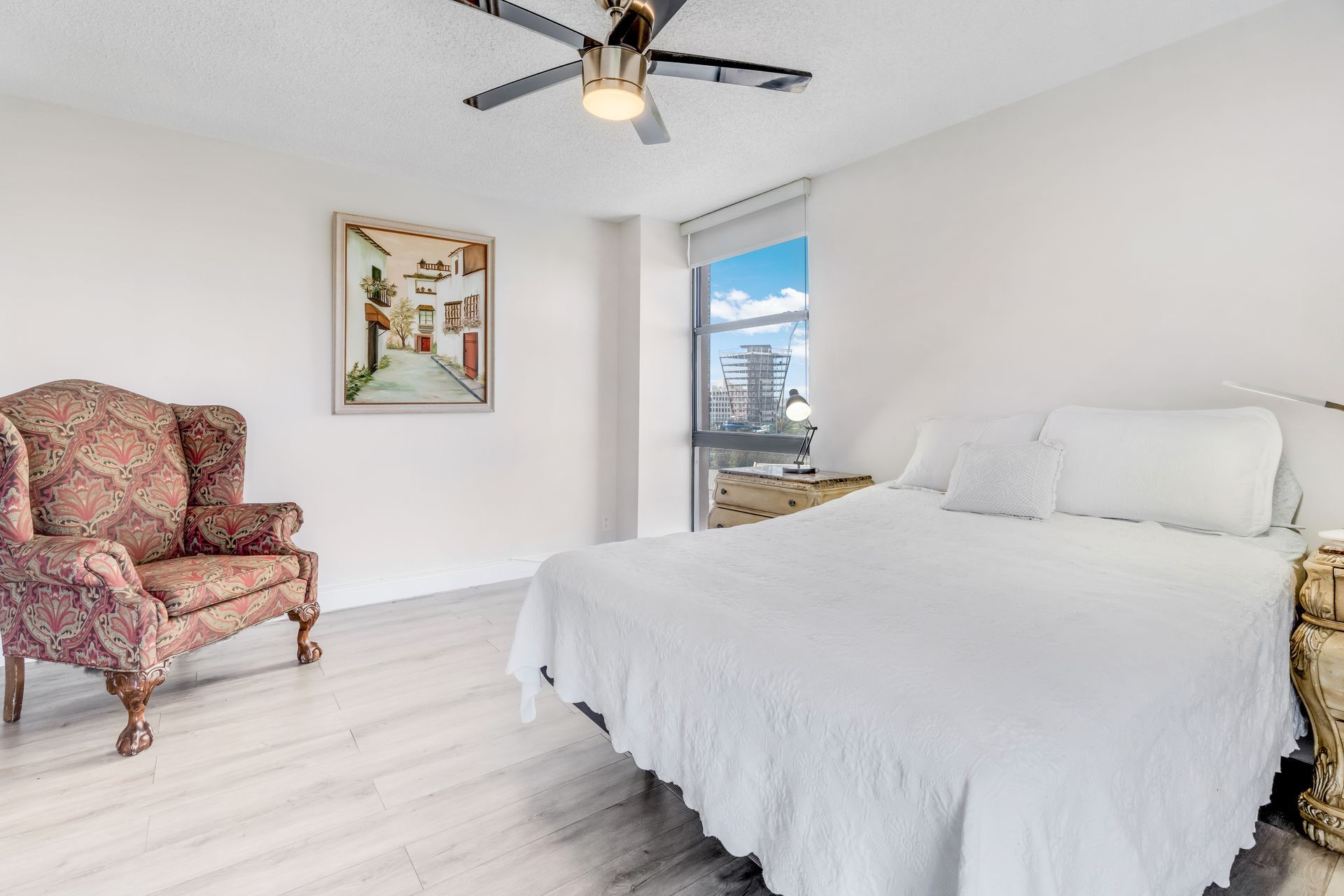 A bedroom with a white-covered bed, a patterned wingback chair, framed wall art, and a large window with city views.