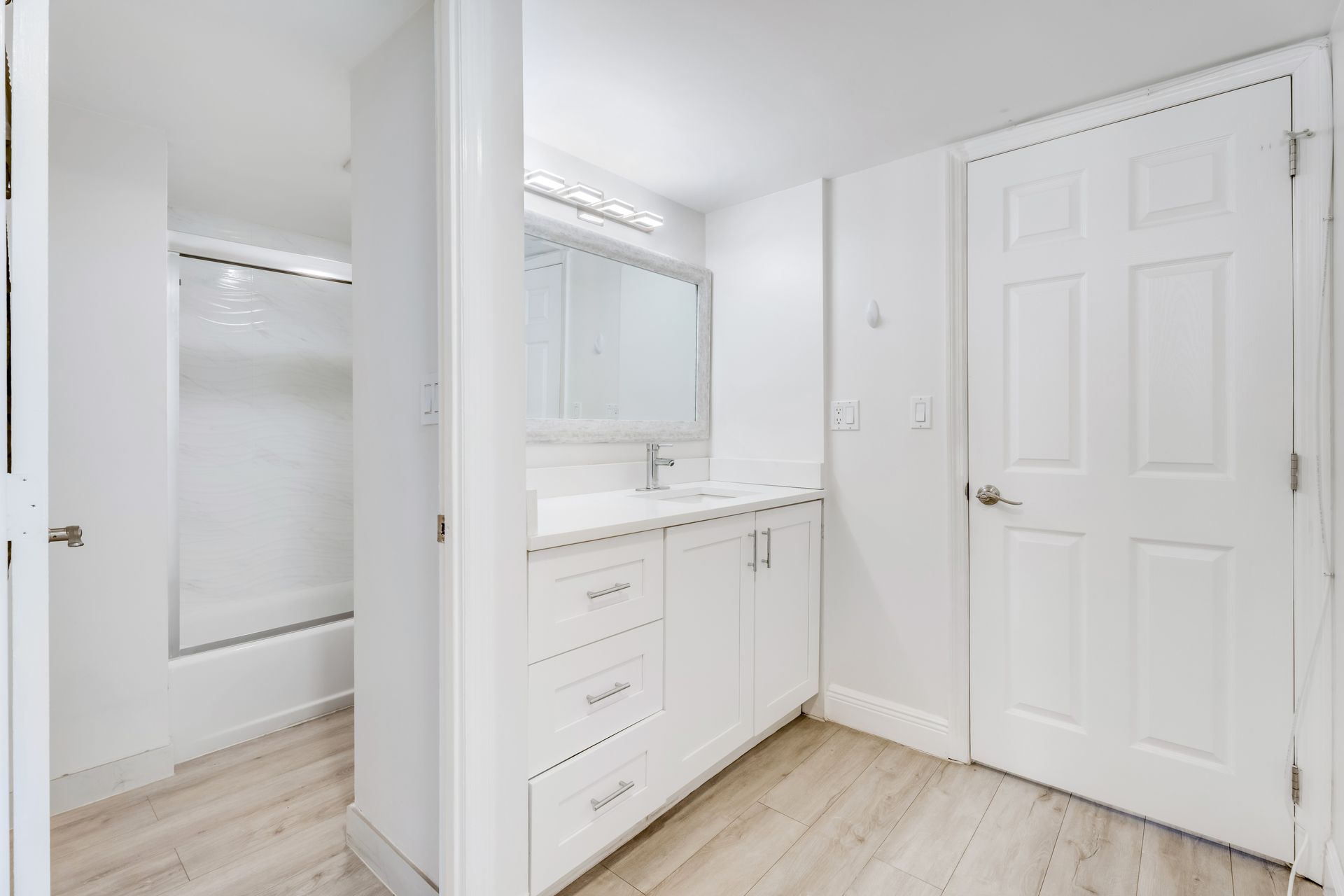 A modern, all-white bathroom featuring a vanity with drawers, a wall-mounted mirror, light wood floors, and a walk-in shower.