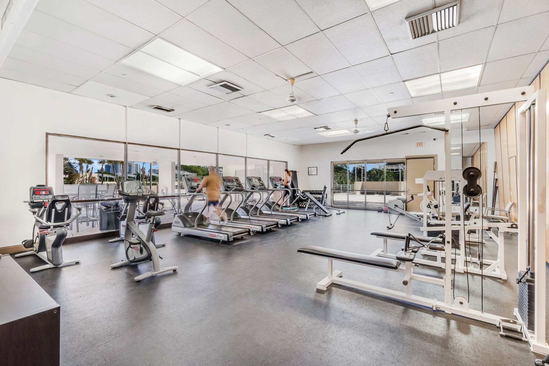 A bright gym with a mirror-lined wall, rows of treadmills, exercise bikes, and strength-training equipment.