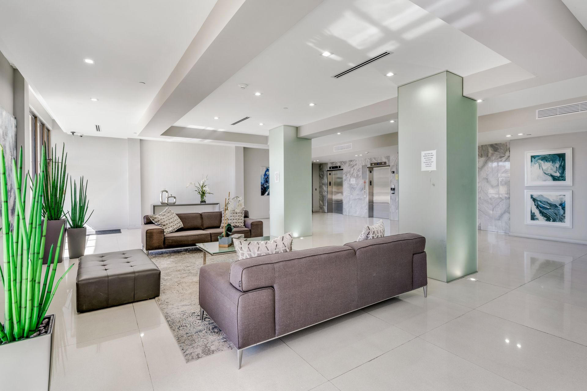 A modern, bright building lobby with two couches, an ottoman, a patterned rug, and tall green plants.