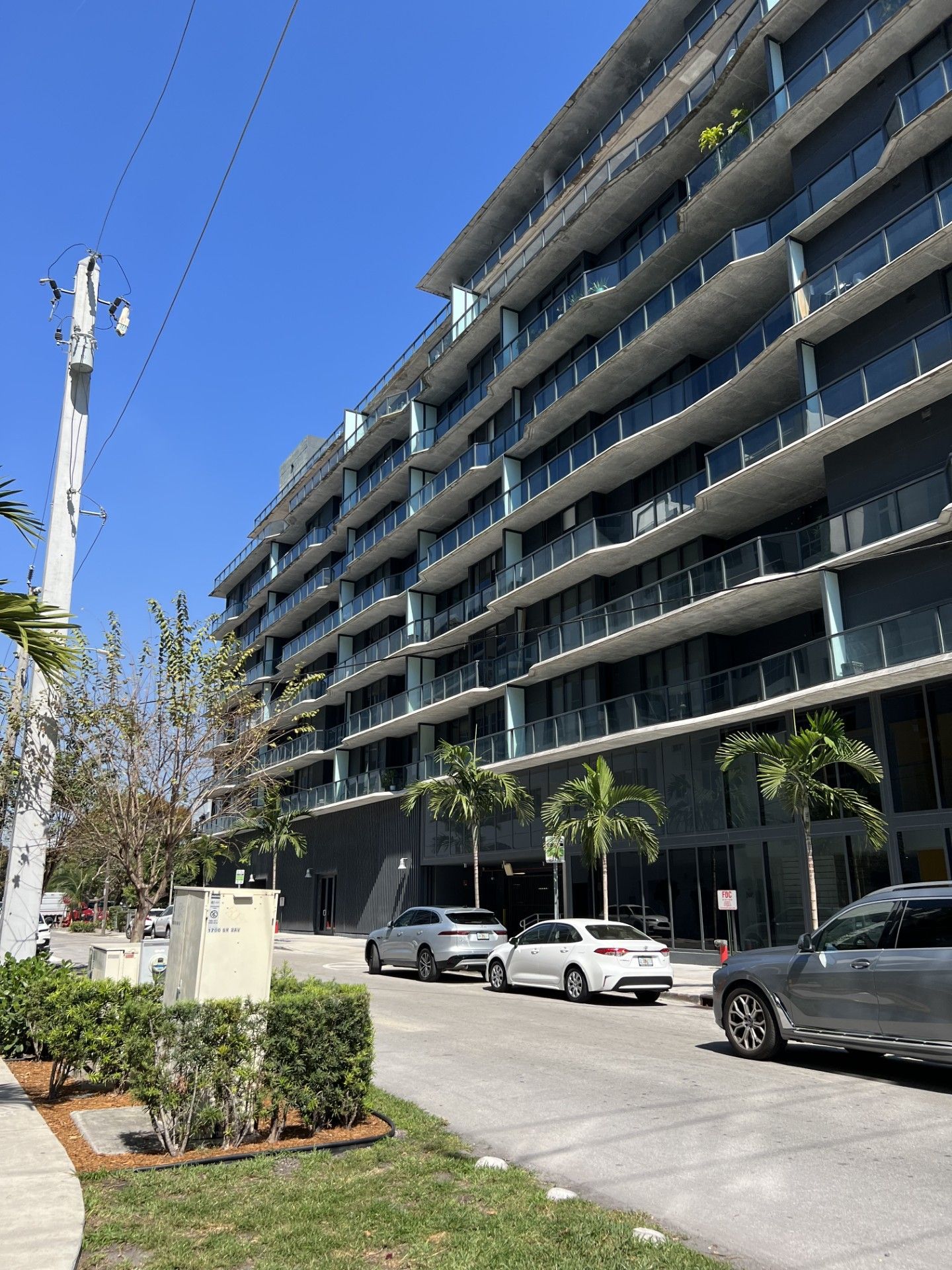 Modern apartment building with glass balconies, parked cars, and palm trees under a bright blue sky.
