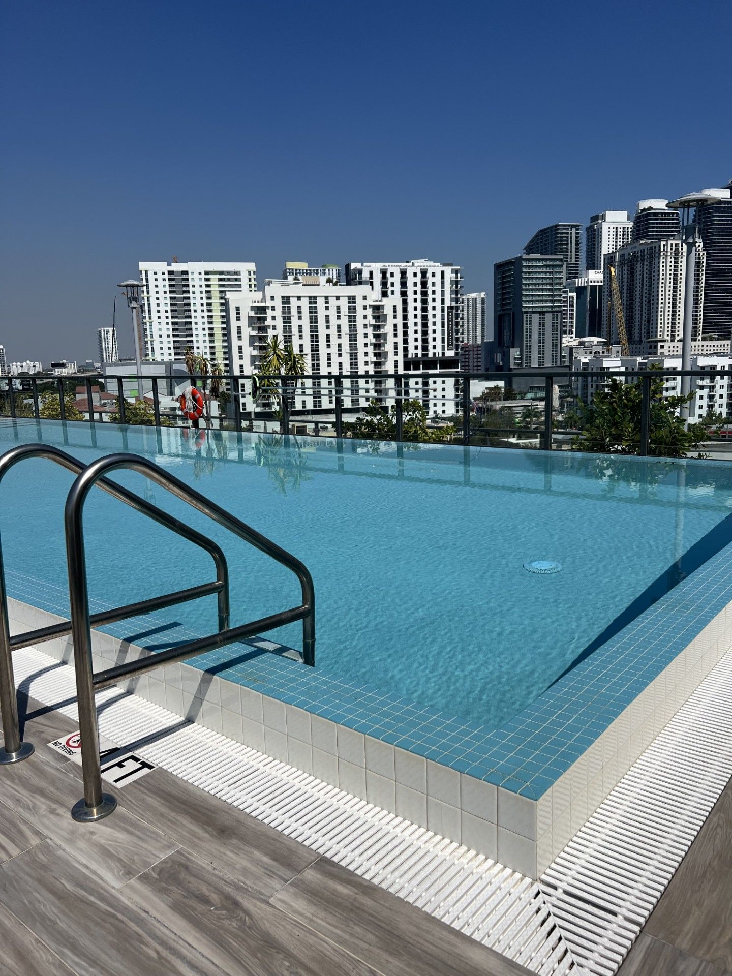 An infinity pool with blue tiled water and a metal ladder, overlooking a city with modern white and grey high-rise towers.