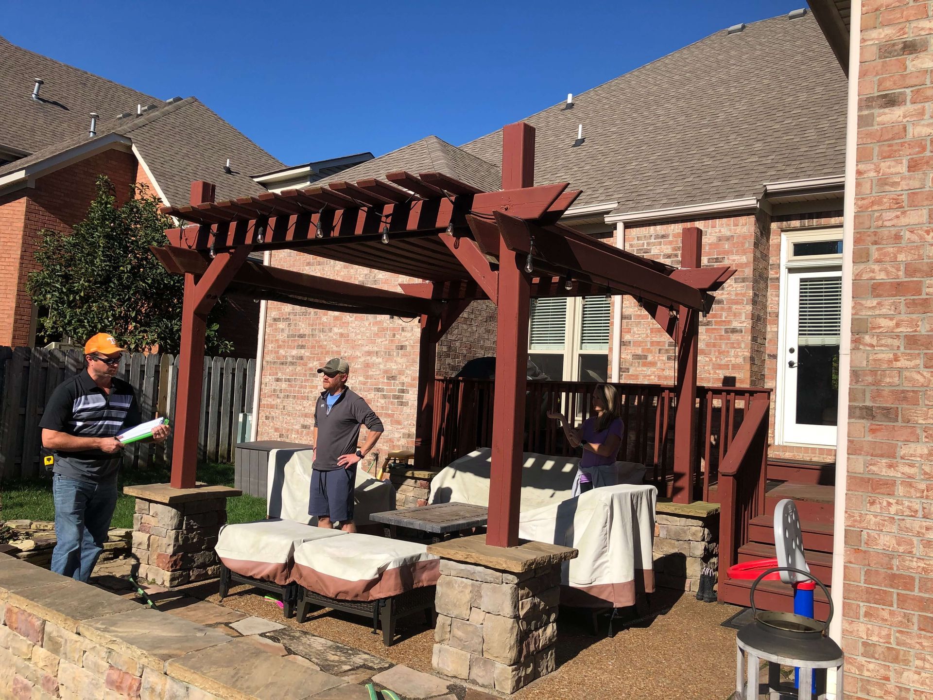 Two men are standing under a pergola in front of a brick house.