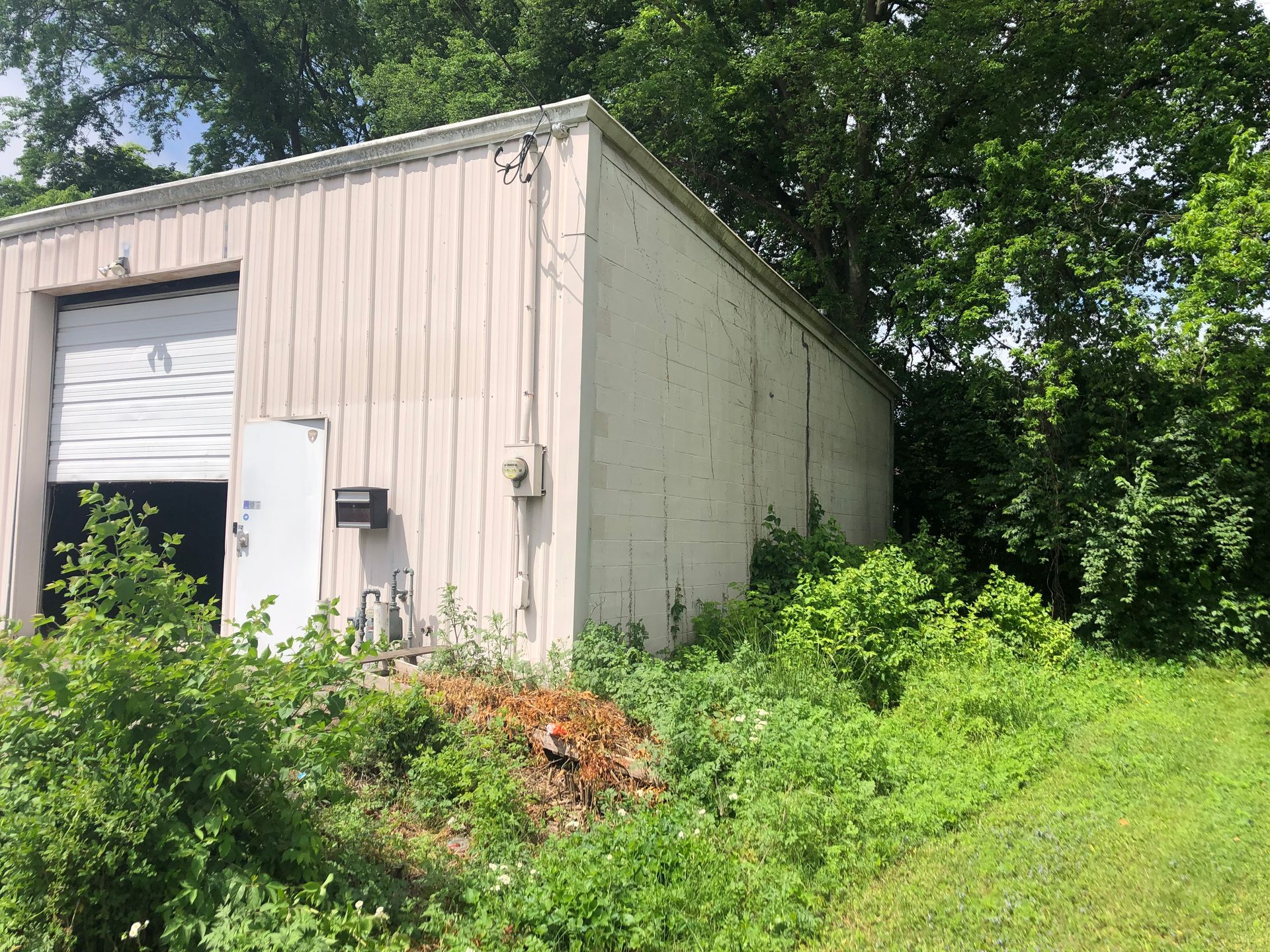 A small white building with a garage door is surrounded by trees and grass.
