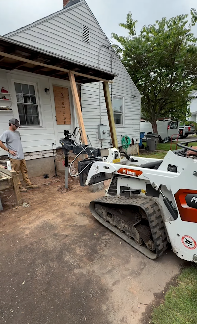 Bobcat tractor and man working on porch renovation. Exterior of house, lumber, and dirt are visible.