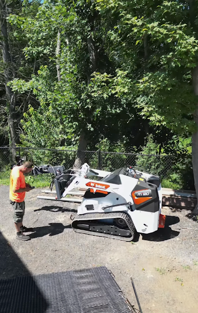Man operates a white and orange Bobcat skid-steer loader outdoors near trees on a sunny day.
