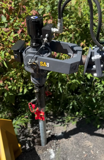 Hydraulic post driver installing a metal pole into the ground, with greenery in the background.