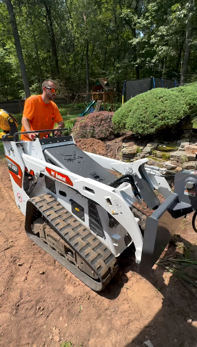 Man in orange shirt operating a white Bobcat track loader in a yard with greenery.