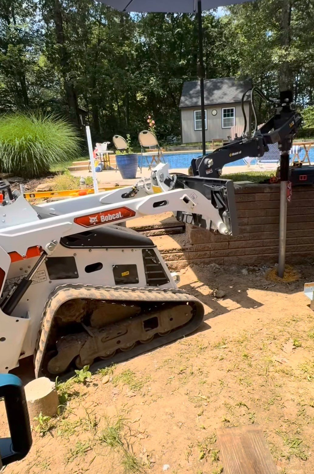 Bobcat skid-steer with auger drilling near a pool in a backyard. A building and umbrella are in the background.