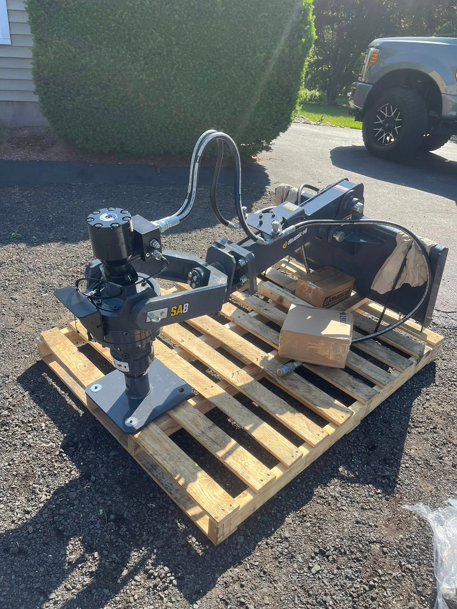 Robotic arm on wooden pallet, outdoors. Gray machine, sunlight, a truck in background.