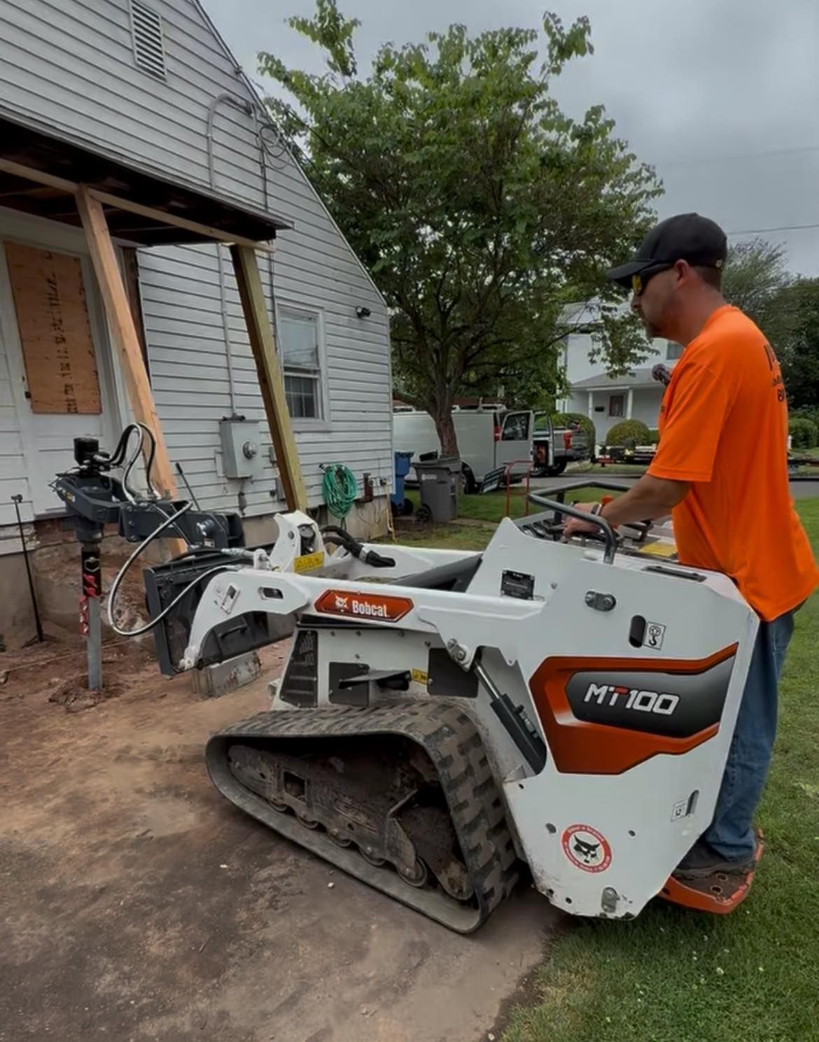 Man in orange shirt operating a white Bobcat MT100 skid-steer loader next to a house under construction.