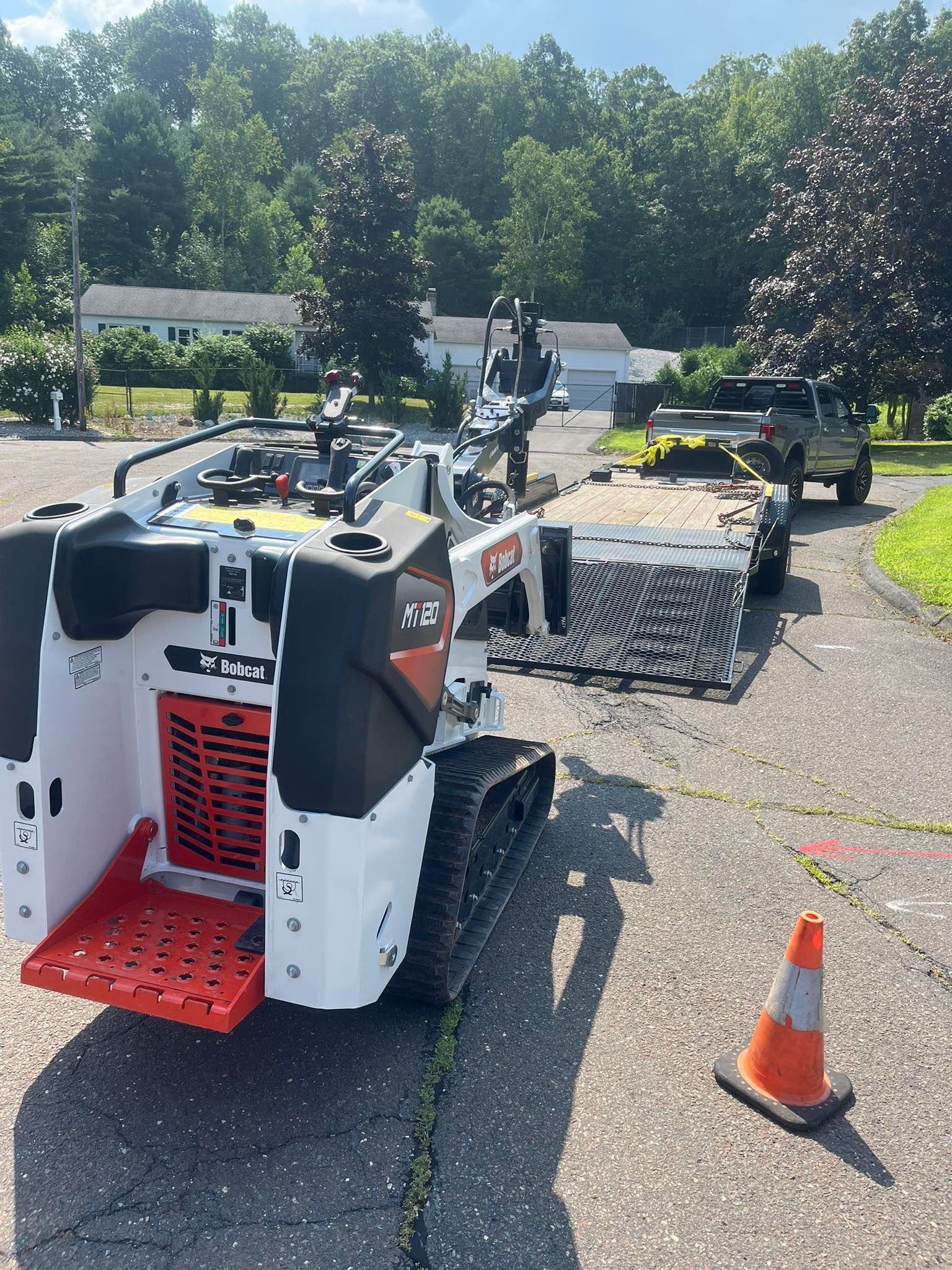 Bobcat Z45 excavator on tracks with trailer, orange cone on asphalt path, green trees, sunny day.