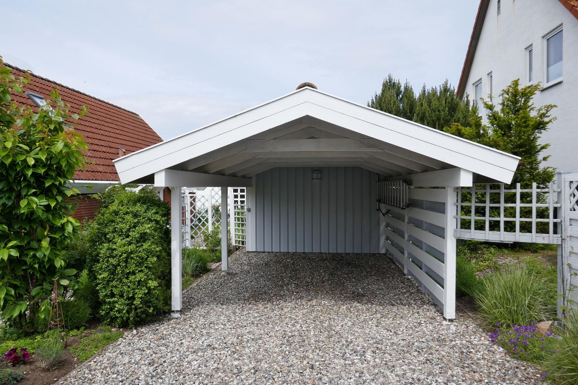 White carport with gravel driveway; light blue walls, surrounded by greenery and houses.
