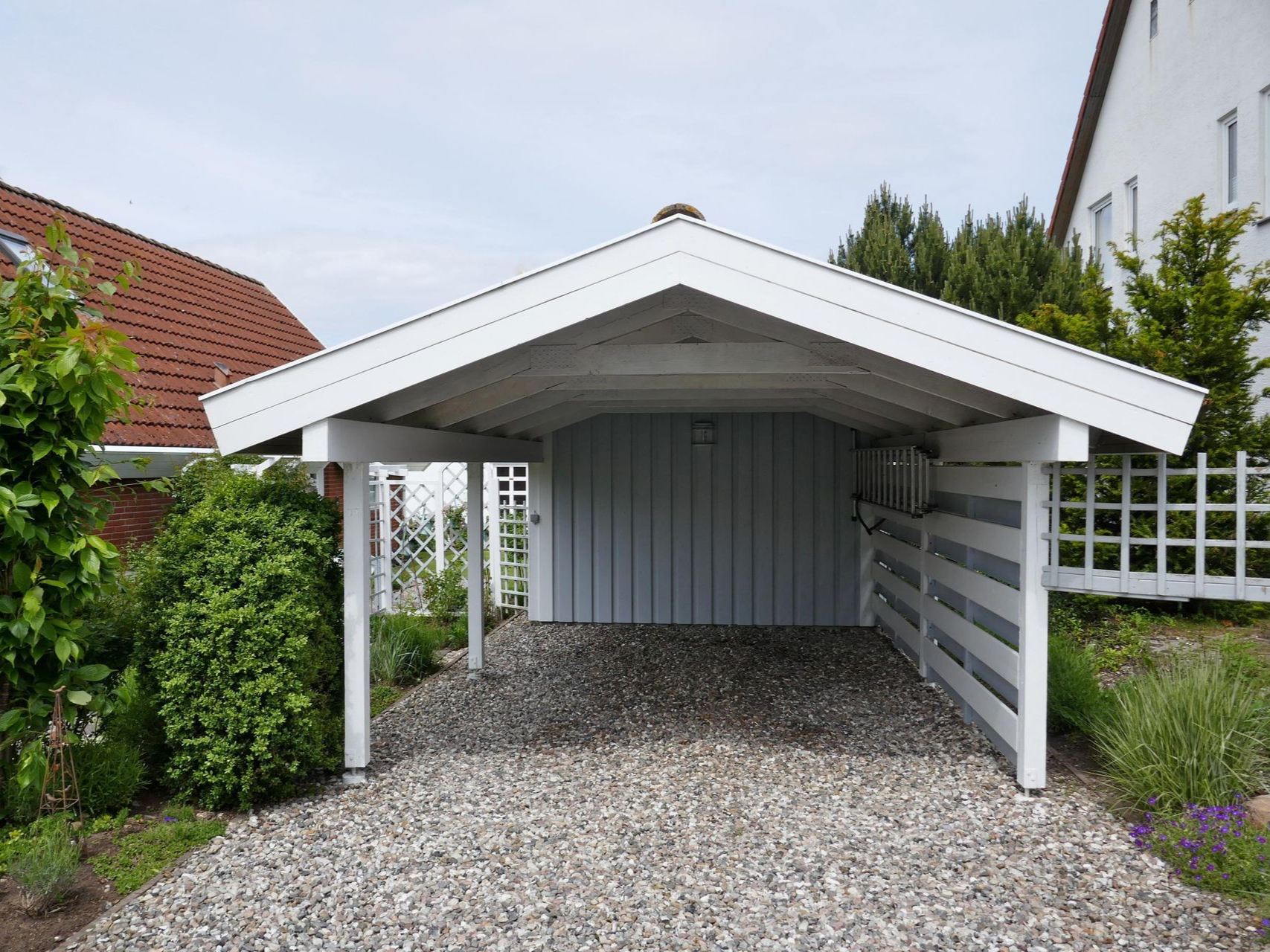 White carport with gray gravel driveway, blue-gray back wall, and white picket fence in a residential setting.