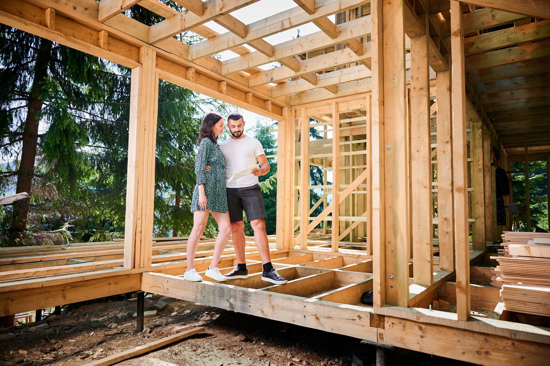 Couple standing in the wooden frame of a house under construction, looking at plans.
