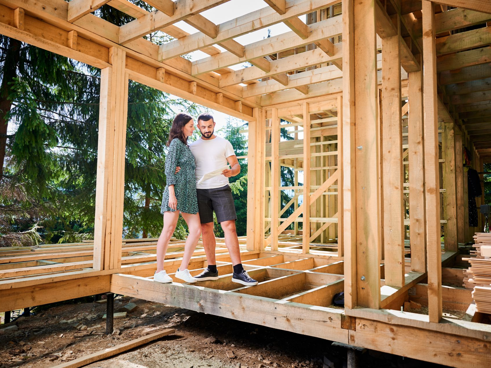 Couple standing inside wooden house under construction, looking at plans.