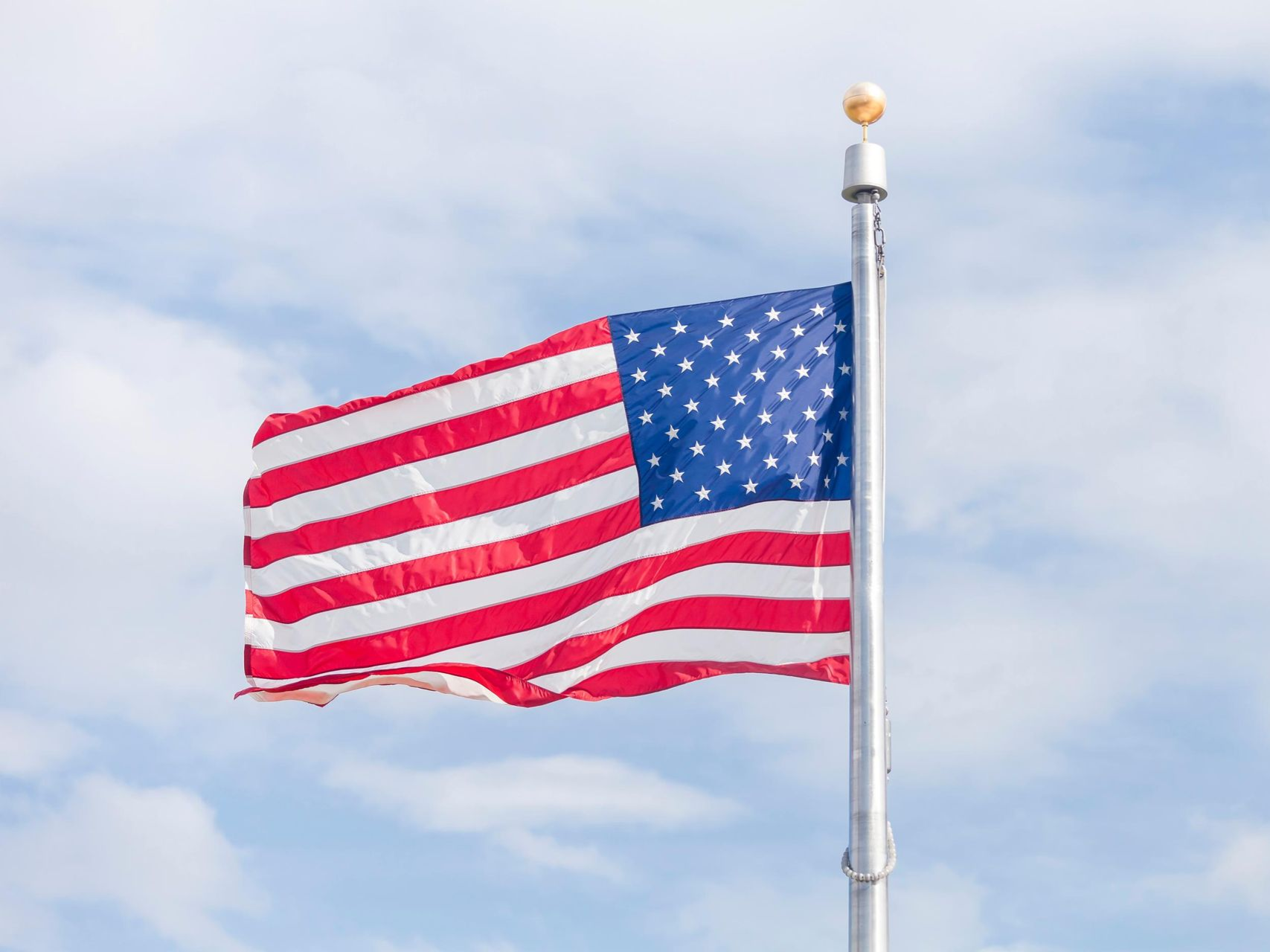 American flag waving in the wind against a cloudy blue sky, atop a silver flagpole.