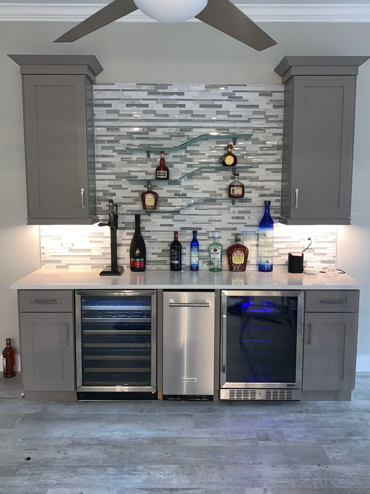 Gray built-in bar with cabinets, appliances, bottles, and a mosaic backsplash.