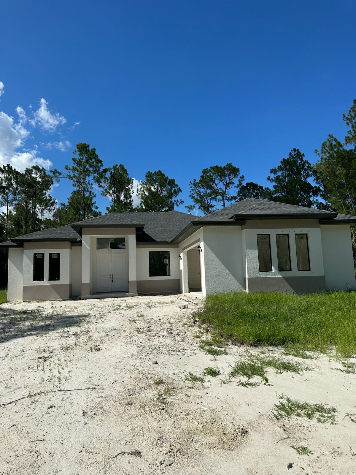 New light-colored house with dark roof and trim under a blue sky, on a dirt lot with some green grass.