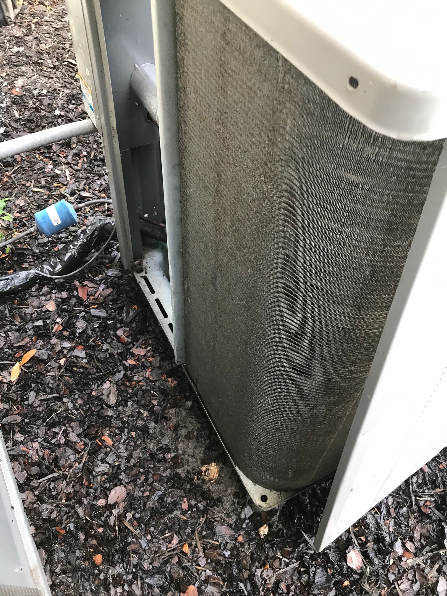 Outdoor air conditioner unit showing a dirty, dark gray condenser coil sitting on a mulch-covered ground.